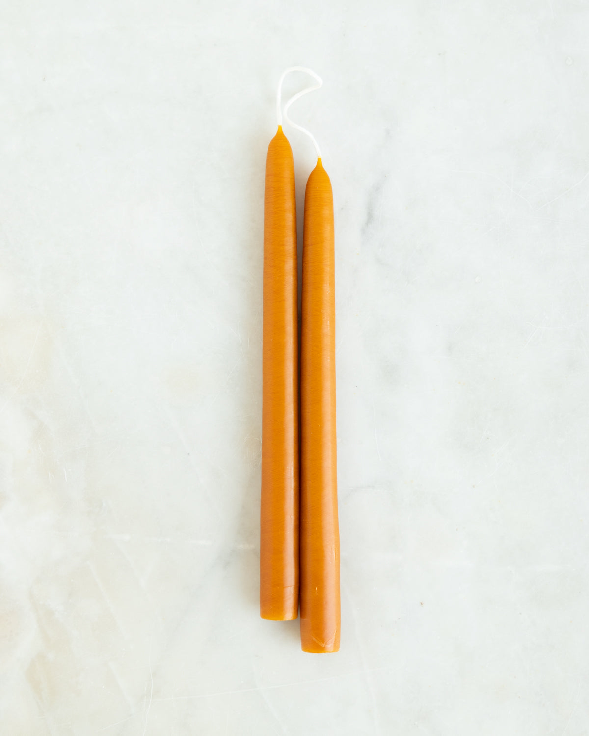 A pair of hand dipped creamy orange taper candles on a lightly marbled white background.