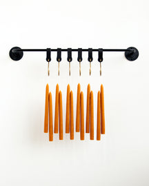 A dozen orange taper candles hanging in pairs from black leather hooks on a black wall pot rack.